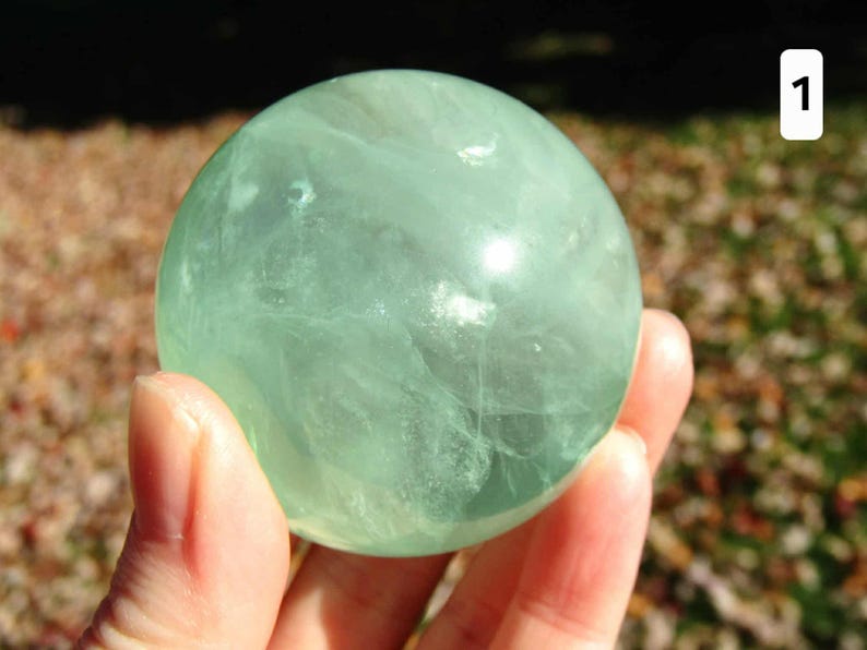 May include: A translucent, light green fluorite sphere is held in a hand. The crystal ball has internal veining and a smooth, polished surface. The background is blurred, with hints of brown and green.