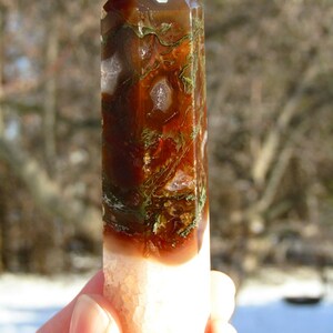May include: A polished crystal obelisk with a faceted top. The stone features a mix of brown, green, and white colors. The bottom portion is a light peach color. The obelisk is held in a hand.