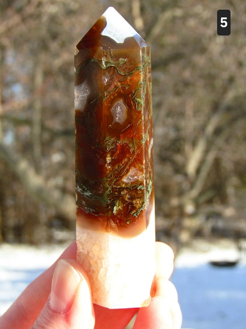 May include: A polished crystal obelisk with a faceted top. The stone features a mix of brown, green, and white colors. The bottom portion is a light peach color. The obelisk is held in a hand.