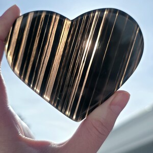 May include: A heart-shaped, polished stone with a striped pattern. The stone is dark, with light brown and black stripes. The heart is held up against a bright, sunny sky. The stone appears to be translucent, with light shining through the stripes.