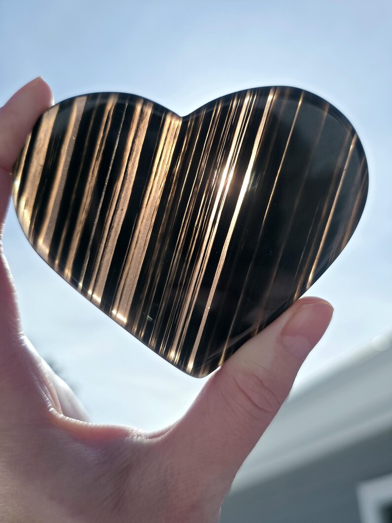 May include: A heart-shaped, polished stone with a striped pattern. The stone is dark, with light brown and black stripes. The heart is held up against a bright, sunny sky. The stone appears to be translucent, with light shining through the stripes.