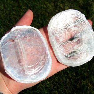 Op de afbeelding: Twee cirkelvormige, doorschijnende witte selenietkristallen met een vezelige, gelaagde textuur. De stenen vertonen een natuurlijke, reflecterende glans en een gladde, licht onregelmatige rand, die de karakteristieke mineraalstructuur van gips laat zien.
