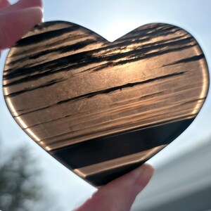 May include: A heart-shaped stone with a translucent, smoky brown appearance featuring distinct horizontal black bands and inclusions. The surface is smooth and polished, allowing light to pass through the semi-transparent material.