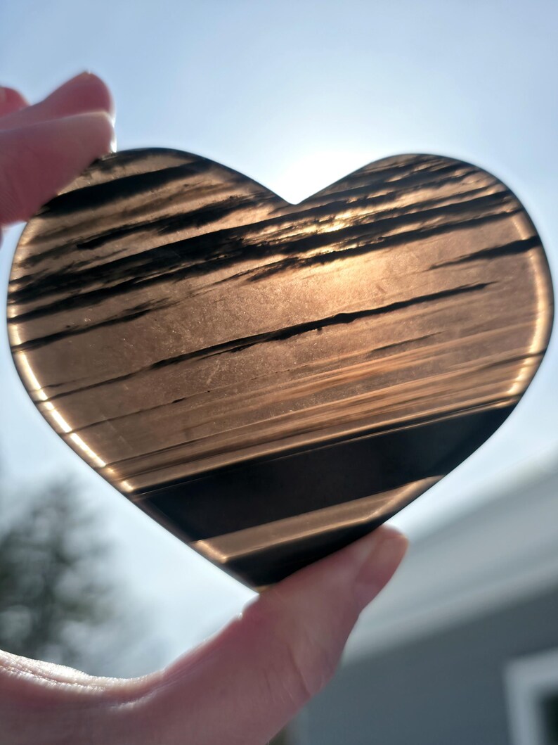 May include: A heart-shaped stone with a translucent, smoky brown appearance featuring distinct horizontal black bands and inclusions. The surface is smooth and polished, allowing light to pass through the semi-transparent material.