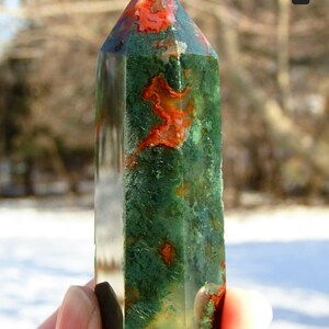 May include: A polished gemstone obelisk featuring a mix of green and red hues. The obelisk has a pointed top and a rectangular base. The stone's surface has a natural, textured appearance. The obelisk is held in a person's hand.