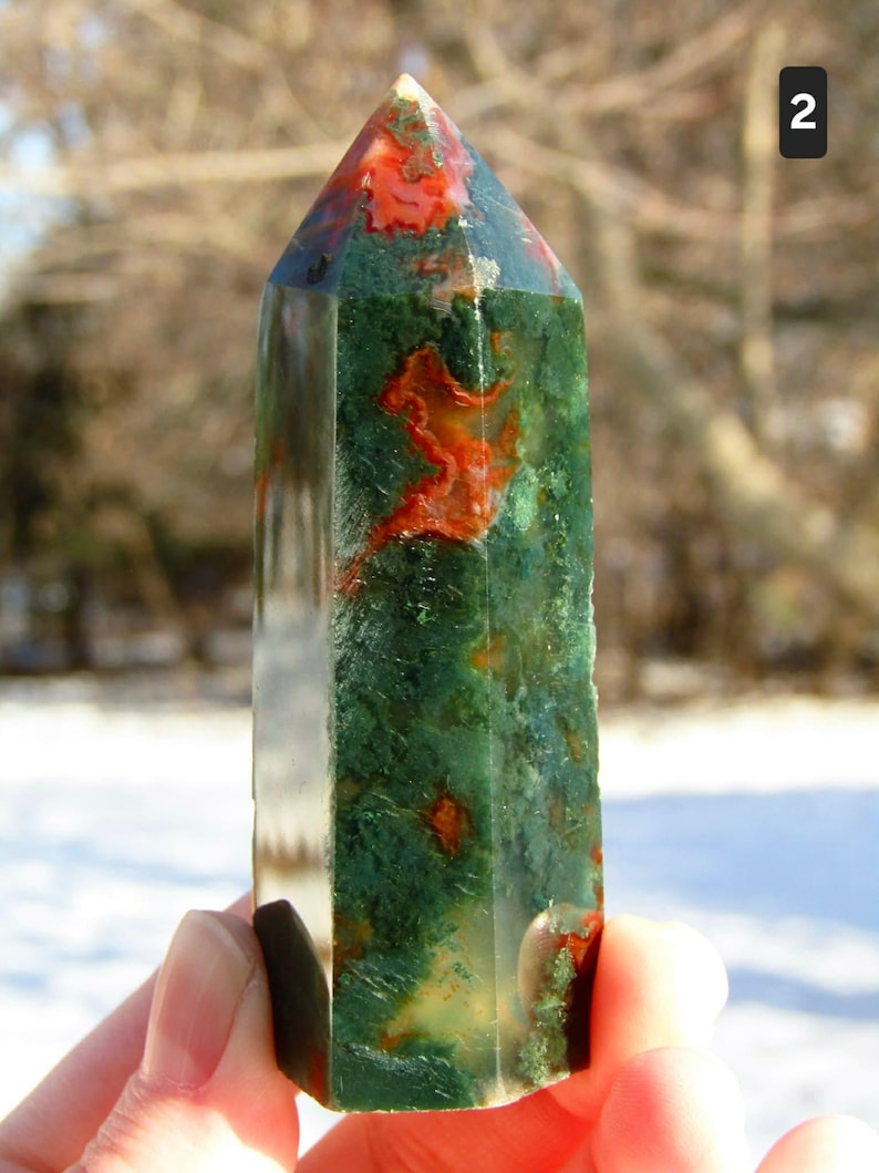 May include: A polished gemstone obelisk featuring a mix of green and red hues. The obelisk has a pointed top and a rectangular base. The stone's surface has a natural, textured appearance. The obelisk is held in a person's hand.