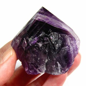 May include: A large, purple amethyst crystal with a pointed top and a rough, natural texture. The crystal is held in a hand.