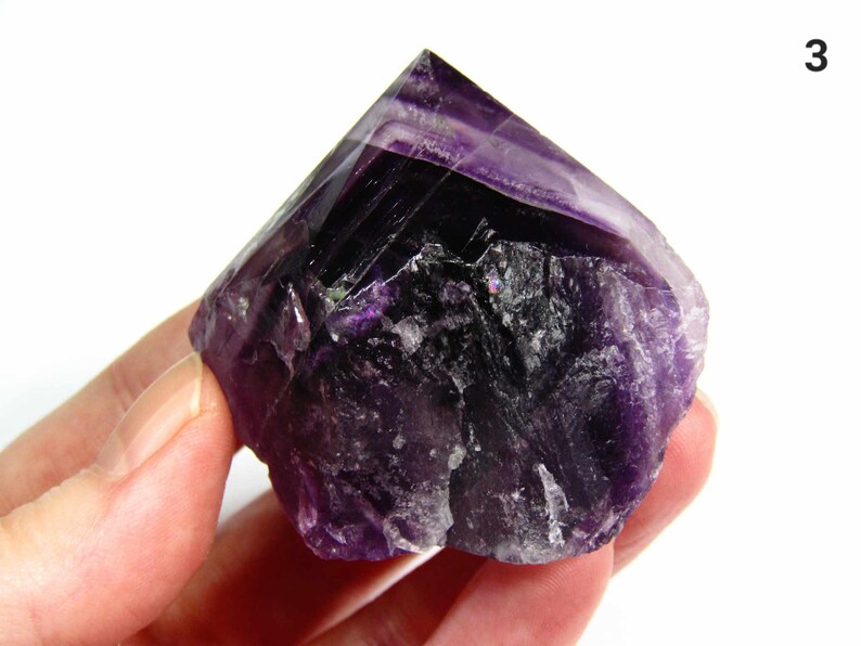 May include: A large, purple amethyst crystal with a pointed top and a rough, natural texture. The crystal is held in a hand.