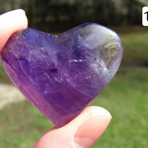 Puede incluir: Un cristal de amatista en forma de coraz&oacute;n, sostenido entre los dedos, muestra un tono p&uacute;rpura intenso con &aacute;reas transl&uacute;cidas. La piedra pulida tiene una superficie lisa, que refleja la luz. El fondo est&aacute; borroso, con el n&uacute;mero blanco 13 en la esquina superior derecha.