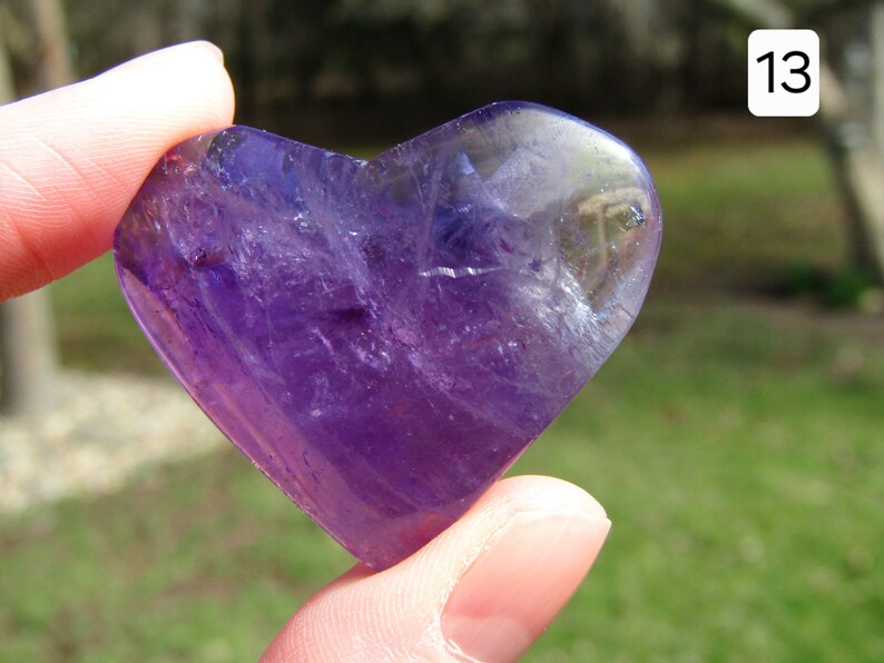 Puede incluir: Un cristal de amatista en forma de coraz&oacute;n, sostenido entre los dedos, muestra un tono p&uacute;rpura intenso con &aacute;reas transl&uacute;cidas. La piedra pulida tiene una superficie lisa, que refleja la luz. El fondo est&aacute; borroso, con el n&uacute;mero blanco 13 en la esquina superior derecha.