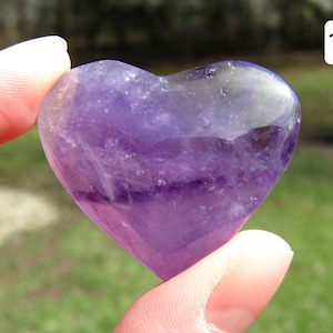 Puede incluir: Una piedra preciosa de amatista en forma de coraz&oacute;n, que muestra tonos de p&uacute;rpura y &aacute;reas transl&uacute;cidas. La piedra pulida se sostiene entre dos dedos, con un fondo verde borroso.
