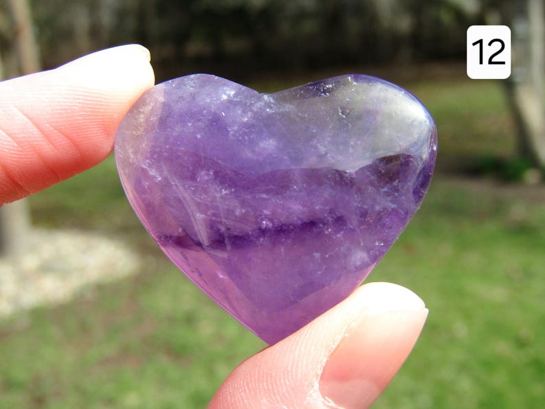 Puede incluir: Una piedra preciosa de amatista en forma de coraz&oacute;n, que muestra tonos de p&uacute;rpura y &aacute;reas transl&uacute;cidas. La piedra pulida se sostiene entre dos dedos, con un fondo verde borroso.