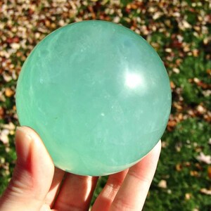 May include: A translucent, light green sphere, held in a hand, against a blurred background of green grass and fallen leaves. The sphere has a smooth, polished surface. A white number 4 is in the upper right corner.