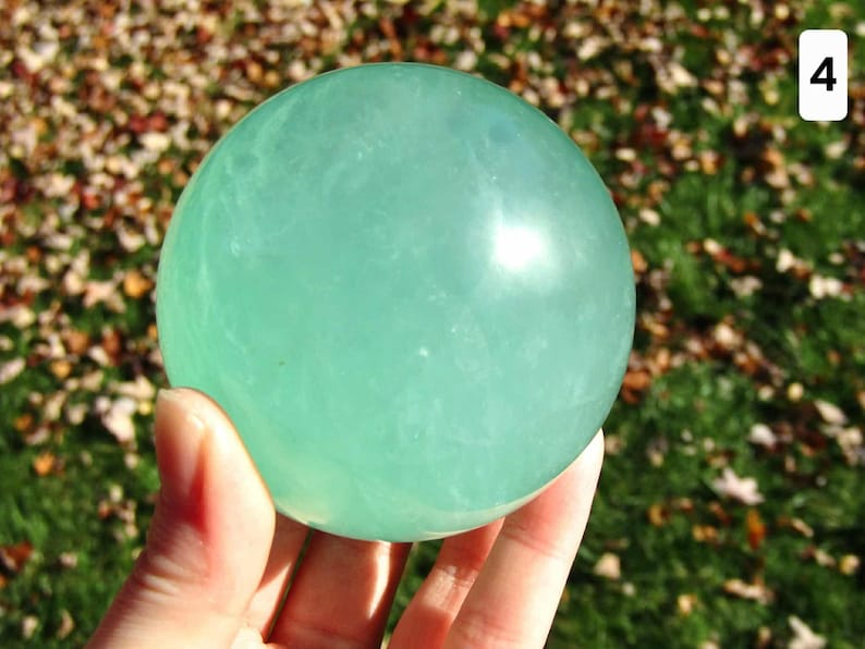 May include: A translucent, light green sphere, held in a hand, against a blurred background of green grass and fallen leaves. The sphere has a smooth, polished surface. A white number 4 is in the upper right corner.