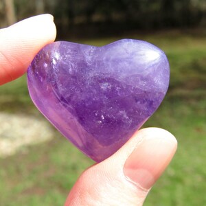Puede incluir: Una piedra preciosa de amatista en forma de coraz&oacute;n, sostenida entre los dedos. La piedra pulida muestra un tono p&uacute;rpura intenso con vetas m&aacute;s claras, reflejando la luz. La forma de coraz&oacute;n es suave y sim&eacute;trica, sugiriendo curaci&oacute;n y amor.