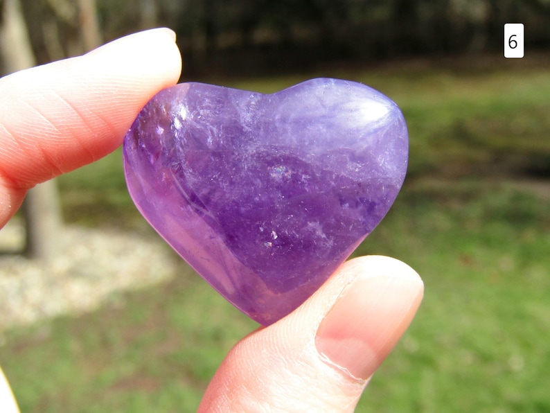 Puede incluir: Una piedra preciosa de amatista en forma de coraz&oacute;n, sostenida entre los dedos. La piedra pulida muestra un tono p&uacute;rpura intenso con vetas m&aacute;s claras, reflejando la luz. La forma de coraz&oacute;n es suave y sim&eacute;trica, sugiriendo curaci&oacute;n y amor.