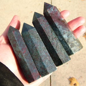 May include: Four polished gemstone obelisks in shades of teal and burgundy are held in a hand. The obelisks have a faceted design and are of varying lengths, showcasing natural stone patterns.