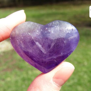 Puede incluir: Una piedra preciosa de amatista en forma de coraz&oacute;n, que muestra tonos de p&uacute;rpura y lavanda. La piedra pulida se sostiene entre los dedos, mostrando su superficie lisa y sus variaciones de color naturales. El n&uacute;mero "16" est&aacute; en la esquina superior derecha.