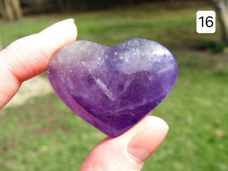Puede incluir: Una piedra preciosa de amatista en forma de coraz&oacute;n, que muestra tonos de p&uacute;rpura y lavanda. La piedra pulida se sostiene entre los dedos, mostrando su superficie lisa y sus variaciones de color naturales. El n&uacute;mero "16" est&aacute; en la esquina superior derecha.