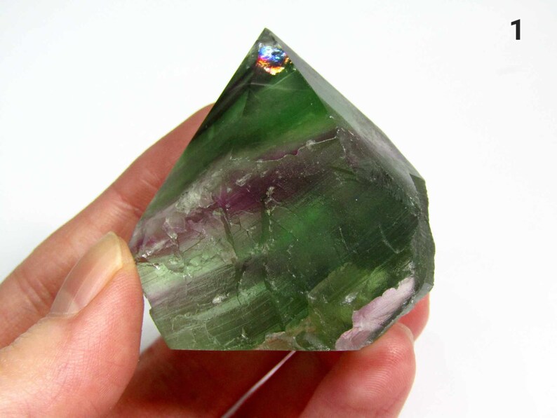 May include: A green, purple, and white fluorite crystal point with iridescent facets. The crystal is approximately 2 inches tall and is held in a hand.