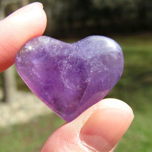 Puede incluir: Una piedra preciosa de amatista pulida en forma de coraz&oacute;n. La piedra p&uacute;rpura se sostiene entre los dedos, mostrando su superficie lisa y reflectante. El color de la piedra var&iacute;a de violeta claro a oscuro.