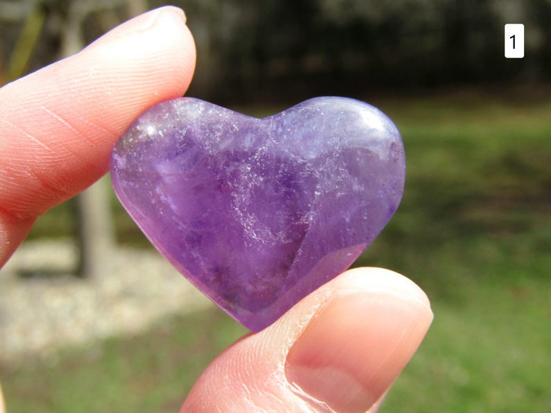 Puede incluir: Una piedra preciosa de amatista pulida en forma de coraz&oacute;n. La piedra p&uacute;rpura se sostiene entre los dedos, mostrando su superficie lisa y reflectante. El color de la piedra var&iacute;a de violeta claro a oscuro.
