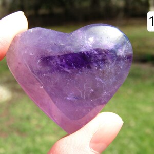 Puede incluir: Un cristal de amatista en forma de coraz&oacute;n, sostenido entre los dedos, muestra tonos de p&uacute;rpura y violeta. La piedra pulida tiene una calidad transl&uacute;cida, con inclusiones naturales visibles. El n&uacute;mero "19" est&aacute; en un cuadro blanco en la esquina superior derecha.