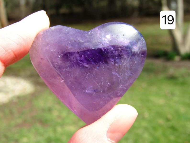 Puede incluir: Un cristal de amatista en forma de coraz&oacute;n, sostenido entre los dedos, muestra tonos de p&uacute;rpura y violeta. La piedra pulida tiene una calidad transl&uacute;cida, con inclusiones naturales visibles. El n&uacute;mero "19" est&aacute; en un cuadro blanco en la esquina superior derecha.