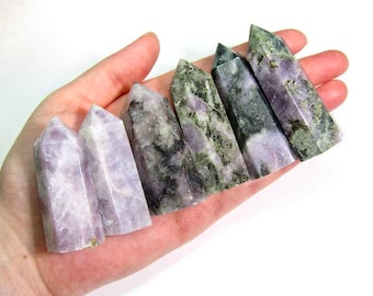May include: Seven purple and green amethyst crystal points, also known as amethyst scepters, are arranged in a row on a hand. The points are various sizes and have a natural, rough texture.