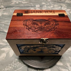 May include: A rectangular wooden box with a reddish-brown lid featuring an engraved eagle design and the text THANK YOU. The box has black metal hinges, a silver-colored front latch, and a light-colored wooden frame surrounding a blue panel.