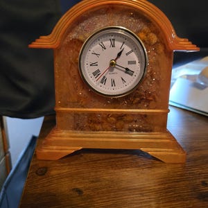 May include: An arched mantel clock made of translucent orange resin with embedded dried flowers and glitter. The clock face is white with black Roman numerals, black hour and minute hands, a red second hand, and the word Quartz printed in black.