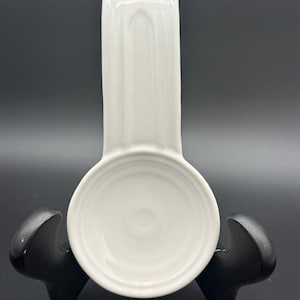 May include: A white ceramic spoon rest featuring a circular basin and an elongated, curved handle section. The surface is smooth and glossy, with a simple, functional design intended to hold kitchen utensils while cooking on a stovetop.