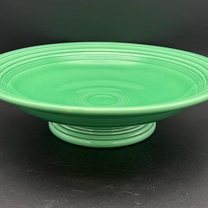 May include: A bright green ceramic pedestal bowl featuring a wide, shallow basin with concentric ring detailing along the rim and the base. The bowl has a smooth, glossy finish and a raised circular foot that elevates the main serving area.