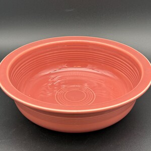 May include: A coral-colored ceramic bowl with a wide rim and a circular, ridged design. The bowl has a smooth, glossy finish and is likely used for serving food. The bowl's diameter is approximately 9 inches.