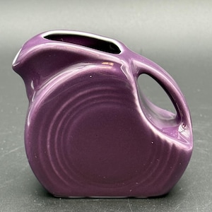May include: A small, purple ceramic pitcher with a handle and a spout. The pitcher has a glossy finish and a rounded body with concentric circle detailing. The interior rim is white.