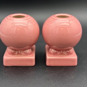 May include: Two matching pink ceramic candle holders. Each has a spherical top with a central opening and sits on a square base with a decorative design. The candle holders are a muted pink color.