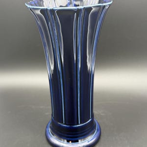 May include: A deep navy blue ceramic vase featuring a vertical ribbed or fluted design that extends from the flared rim down to the circular base. The glossy glaze reflects light along the ridges, highlighting the vertical lines of the structure.