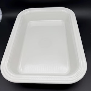 May include: A rectangular, white ceramic baking dish with rounded corners. The dish has a slightly raised rim and a subtle concentric circle design in the center. The overall design is simple and clean.