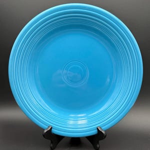May include: A vibrant turquoise ceramic plate with concentric ridged detailing. The plate has a central recessed area and is displayed on a black stand. The plate's bright color and design make it a striking piece for kitchenware.