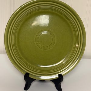 May include: A round, olive green ceramic plate featuring a series of concentric circular ridges along the rim and a smooth, glossy finish. The plate is displayed upright on a black plastic stand, showcasing its uniform color and circular design.