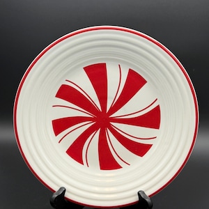 May include: A round white ceramic plate featuring a red peppermint swirl pattern in the center and a thin red band along the outer rim. The plate has a series of concentric circular ridges embossed into the surface, creating a textured appearance.