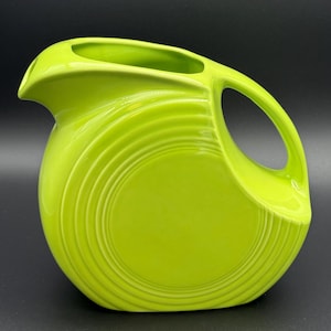 May include: A bright lime green ceramic pitcher with a rounded body and a large handle. The pitcher has a spout for pouring and a series of concentric rings around the base. The pitcher is a vibrant, solid color.