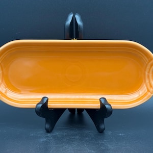 May include: An orange-yellow rectangular serving dish with rounded ends. The dish is made of ceramic and has a glossy finish. It is displayed on a black stand against a dark background. The dish is approximately 12 inches long.