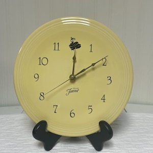 May include: A pale yellow Fiesta plate repurposed as a clock. The clock face features black numerals and hands, with the word "Fiesta" printed below the center. The clock is displayed on a black stand.