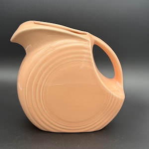 May include: A peach-colored ceramic pitcher with a curved handle and a spout. The pitcher has a rounded body with concentric, ridged detailing. The handle is a simple, curved design. The pitcher is set against a dark background.