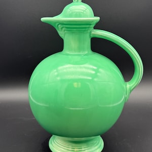 May include: A bright green ceramic pitcher with a spherical body, a narrow neck, a curved handle, and a matching lid. The piece features a glossy finish and a circular base with subtle concentric ring detailing around the bottom and the lid.