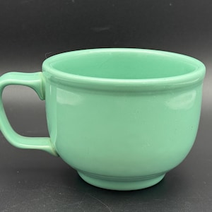 May include: A solid mint green ceramic mug with a rounded bowl shape, a thick rim, and a sturdy C-shaped handle. The mug features a smooth, glossy finish and a slightly raised base, presenting a simple, functional design for holding hot beverages.
