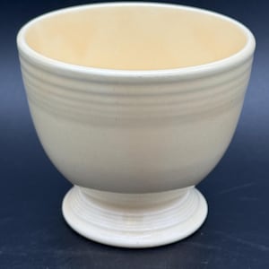 May include: A cream-colored ceramic bowl with a smooth, glossy finish. The bowl features a rounded body with horizontal ribbed detailing near the top rim and a circular pedestal base that also includes subtle horizontal ringed accents.