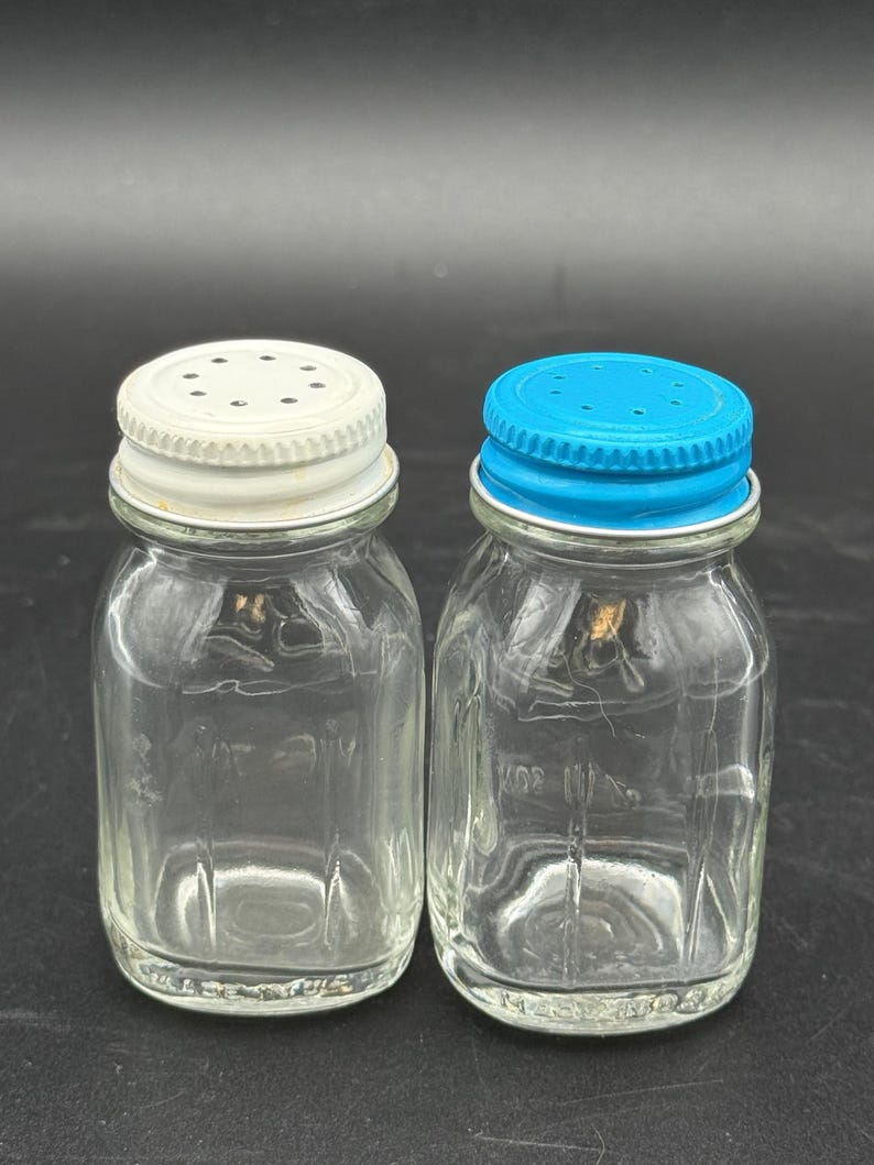 Puede incluir: Dos saleros de vidrio transparente con base cuadrada y esquinas redondeadas. Uno tiene una tapa de rosca de metal blanco con peque&ntilde;os orificios, mientras que el otro tiene una tapa de rosca de metal azul brillante con peque&ntilde;os orificios.
