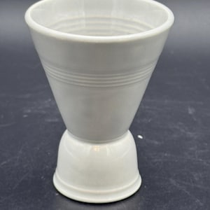 May include: A light gray ceramic jigger with a flared top and a rounded base. The jigger has three horizontal lines around the middle section. The design is simple and elegant, suitable for measuring liquids.